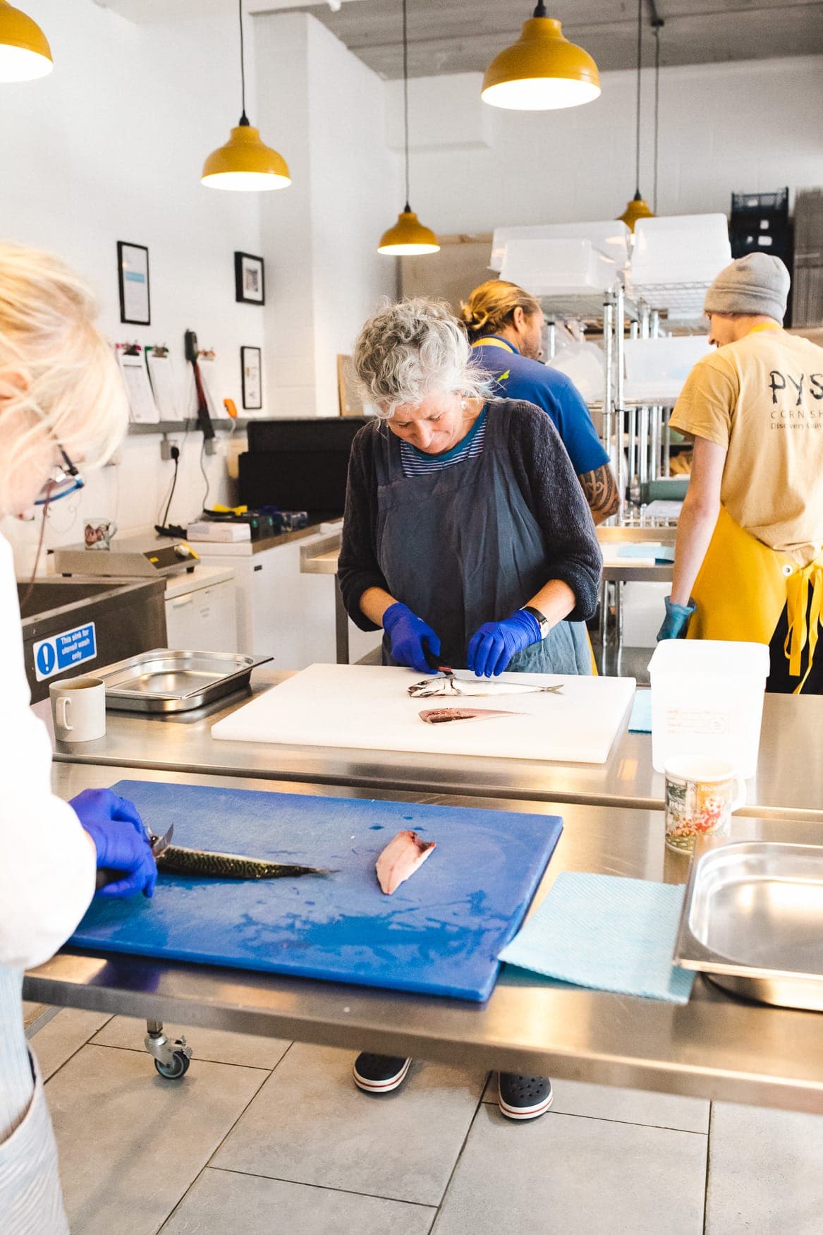 A filleting session at the Pysk shop in Falmouth: several people at stainless steel benches, filleting mackerel on white and blue boards, with a fishmonger in a Pysk apron working alongside them.